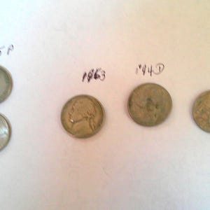 May include: A collection of five vintage nickels, including coins from 1963 and 1944. The image also shows two nickels from 2005. The coins are arranged on a white surface, with handwritten dates above them.