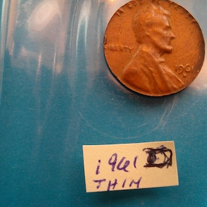 May include: A copper-colored 1941 penny in a protective plastic sleeve. The coin features a profile of a man and the word "LIBERTY". A handwritten note below the coin reads "1941 THIN".