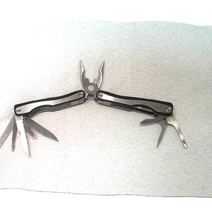 May include: A silver and black multi-tool featuring a pair of pliers at the center, with several folded metal implements extended from the handles, including a serrated blade, a straight blade, and various screwdrivers on both sides.