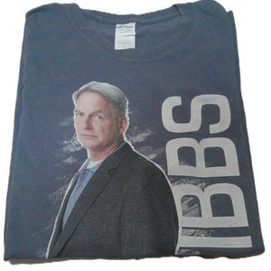 May include: A dark blue short-sleeved t-shirt featuring a photographic print of a man with gray hair wearing a dark suit jacket and a light blue shirt. The text "NCIS" is printed vertically in large, light gray capital letters on the right side.