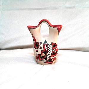May include: A cream-colored ceramic vessel featuring a double-spout design with a central arched handle. The exterior is decorated with geometric patterns in red and black, including triangular shapes, cross-hatching, and circular motifs.