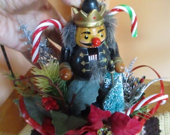 May include: A wooden nutcracker figurine with a gold crown, black jacket, and a red nose. The figurine is surrounded by festive decorations, including candy canes, red berries, and artificial greenery, creating a holiday theme.