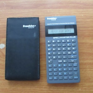 Peut inclure: Un correcteur orthographique portable Franklin Spellmaster Plus noir avec un écran bleu et un clavier avec des lettres, des chiffres et des signes de ponctuation. L'appareil est dans un étui noir avec le logo Franklin Computer.