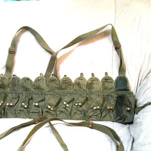 May include: Olive green chest rig with multiple pouches and adjustable straps. The rig features a series of pockets with button closures and a shoulder strap design. The military-style vest is designed to carry ammunition.