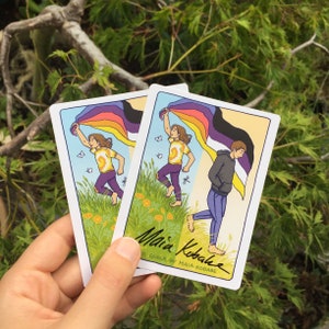 May include: Two illustrated cards featuring people holding a rainbow pride flag. The cards are white with a green, yellow, and orange background. The text on the bottom of the cards reads "Queer by Maia Kobabe".