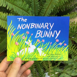 Op de afbeelding: Een rechthoekige boekomslag met de titel The Nonbinary Bunny door Maia Kobabe with deep respect for Margaret Wise Brown and Clement Hurd. De illustratie toont twee witte konijnen in hoog groen gras met kleine roze bloemen.