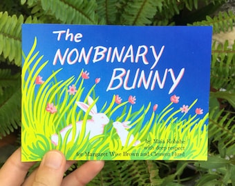 Puede incluir: Una portada de libro rectangular titulada The Nonbinary Bunny por Maia Kobabe with deep respect for Margaret Wise Brown and Clement Hurd. La ilustración muestra dos conejos blancos en hierba verde alta con pequeñas flores rosas.