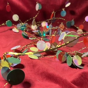 Puede incluir: Una guirnalda decorativa con ramas de alambre dorado y lentejuelas circulares iridiscentes de colores. Las lentejuelas son en tonos de rojo, verde, azul, amarillo y blanco. La guirnalda se exhibe sobre una superficie de terciopelo rojo.