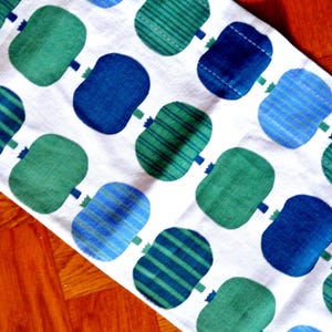 May include: White fabric with a repeating pattern of stylized blue and green shapes. Some shapes have stripes, others are solid. The shapes are connected by small stems. The fabric is likely a table runner or decorative cloth.