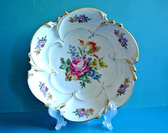 Puede incluir: Una fuente de porcelana blanca con borde festoneado y ribete dorado. La fuente presenta un arreglo floral central con una rosa rosa y otras flores coloridas. Se colocan racimos florales adicionales alrededor del borde. La fuente se exhibe sobre un soporte transparente.