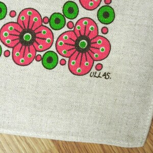 May include: Close-up of a linen textile featuring a repeating floral pattern. The flowers are pink with black centres and green accents. The word "ULLAS" is printed in black in the corner.