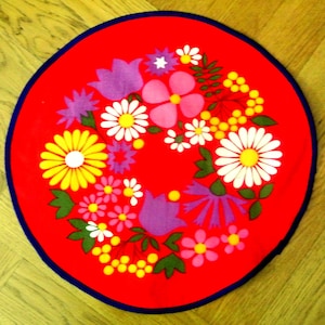 May include: A circular red mat featuring a vibrant floral pattern with white daisies, yellow blossoms, purple tulips, and green leaves. The design is arranged in a wreath-like formation around the center, finished with a dark blue border edge.