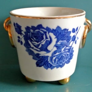 Könnte beinhalten: Ein weißer Keramiktopf mit blauen und goldenen Blumendesigns. Der Topf hat zwei goldene Griffe und drei goldene Füße.