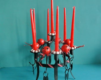 Puede incluir: Un candelabro de metal negro con seis velas rojas y portavelas con forma de cerdo. El candelabro tiene una cinta de rayas blancas y negras alrededor de la base.
