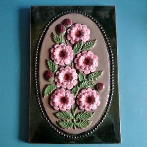 May include: A framed oval-shaped wall decor with a raised floral design. The flowers are pink with dark pink centres and are surrounded by green leaves. The frame is dark green with a silver beaded border.