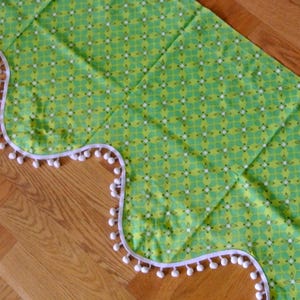 May include: A green and yellow fabric valance with a white pom-pom trim. The valance has a scalloped edge and a repeating geometric pattern.