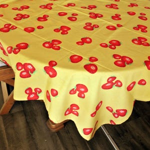 May include: A round yellow tablecloth with a repeating pattern of red strawberries. The tablecloth is draped over a wooden table, with a portion of a chair visible on the left side. The strawberries have green leaves.