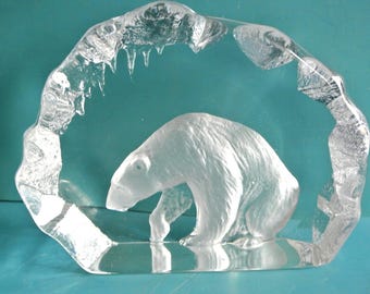 May include: A clear glass sculpture of a polar bear. The bear is shown in a crouching pose, enclosed within a clear, arched frame with icicle details. The sculpture is set against a teal background.