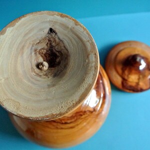 May include: A wooden bowl with a lid, both made of light brown wood with dark brown wood grain patterns. The bowl has a smooth, rounded exterior and a slightly indented interior. The lid has a small knob on top.