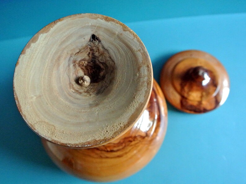 May include: A wooden bowl with a lid, both made of light brown wood with dark brown wood grain patterns. The bowl has a smooth, rounded exterior and a slightly indented interior. The lid has a small knob on top.