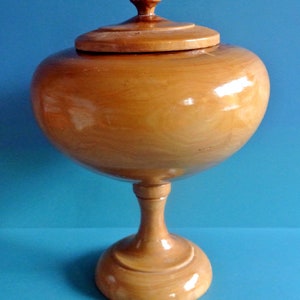 May include: A wooden bowl with a lid, on a pedestal base. The bowl is light brown and has a smooth, polished finish.