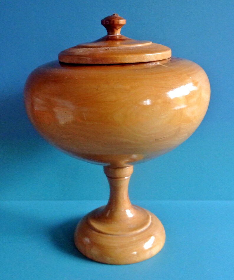 May include: A wooden bowl with a lid, on a pedestal base. The bowl is light brown and has a smooth, polished finish.