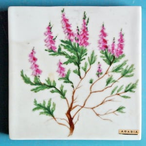 May include: Square ceramic tile with a hand-painted floral design. The artwork features pink flowers, green leaves, and brown branches against a white background. The tile has a small label that reads "ARABIA".