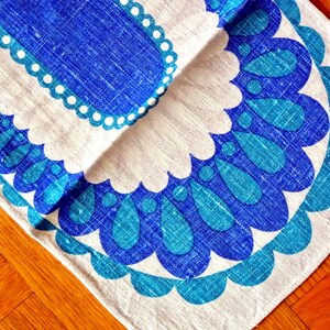 May include: A folded, rectangular textile with a blue and white floral pattern. The design features a central blue shape with a scalloped border, and a larger, stylized floral motif in shades of blue and teal. The fabric appears to be linen or a similar material.