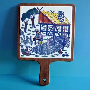Può includere: Un sottopentola in legno con una piastrella in ceramica che presenta un'illustrazione colorata di una fattoria, un uomo e oche. La piastrella è circondata da una cornice in legno e il sottopentola ha un manico in legno.