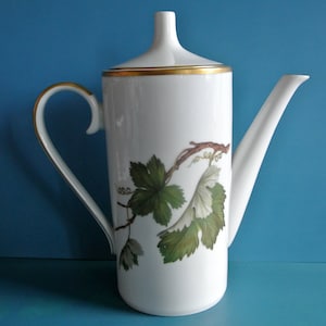 Peut inclure: Cafetière en céramique blanche avec un long bec et une anse incurvée. Le pot présente un bord doré et un motif peint de feuilles et de vignes de raisin vertes et blanches. Le fond est bleu uni.