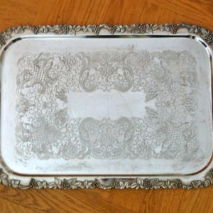 May include: A silver serving tray with ornate, embossed floral designs around the edge and handles. The tray has a rectangular shape and a smooth, reflective surface.