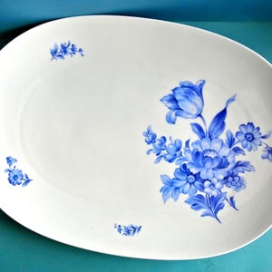May include: White oval serving platter with a blue floral design. The platter features a large cluster of blue flowers on the right side and smaller floral accents on the left. The flowers have detailed petals and leaves.