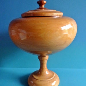 May include: A wooden bowl with a lid, on a pedestal base. The bowl is light brown and has a smooth, polished finish.