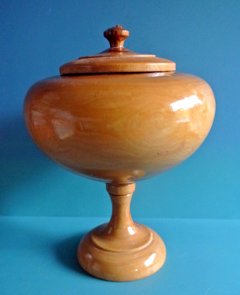 May include: A wooden bowl with a lid, on a pedestal base. The bowl is light brown and has a smooth, polished finish.