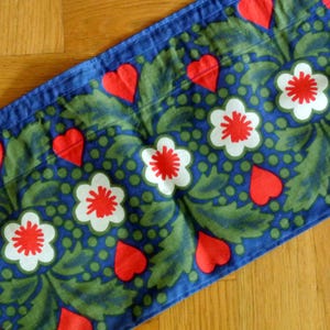 May include: Blue fabric with a repeating pattern of red hearts, white flowers with red centers, and green leaves.