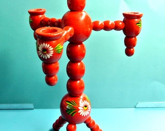 May include: A bright red wooden candle holder featuring a central vertical column made of stacked spheres. It has three arms extending outward, each topped with a candle cup. The base and candle cups are decorated with hand-painted white daisies.