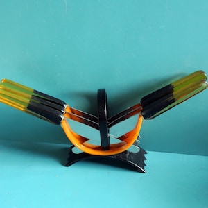 May include: A vintage, abstract fruit basket with a black handle and base, and an orange and black body. The basket has several sections with yellow and green accents, creating a unique, artistic design.