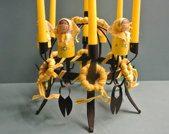 Puede incluir: Un candelabro de metal negro con velas amarillas y muñecas de tela amarillas con caras pintadas. El candelabro tiene una base circular y cuatro brazos.