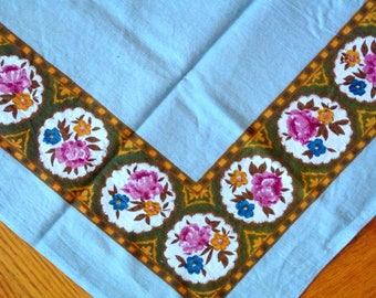 May include: A blue tablecloth with a brown and green floral border. The border features a repeating pattern of pink roses, yellow flowers, and blue leaves.
