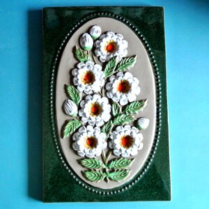 Op de afbeelding: Een rechthoekige keramische tegel met een verhoogd bloemenontwerp. Witte bloemen met oranje centra en groene bladeren tegen een beige ovaal. De tegel heeft een donkergroene rand en een parelrand, waardoor een decoratieve wanddecoratie ontstaat.