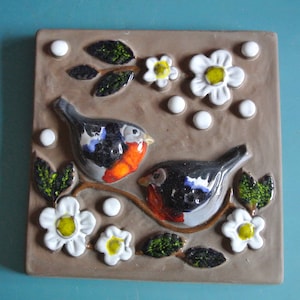 Puede incluir: Un azulejo de cerámica con fondo marrón presenta dos pájaros posados en una rama con flores blancas. Los pájaros son azules, negros, blancos y naranjas. Las flores son blancas con centros amarillos.