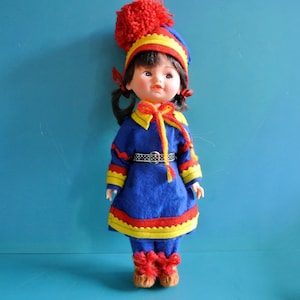 May include: A vintage doll wearing a traditional outfit. The doll has black hair and is dressed in a blue felt coat and pants with yellow and red trim. A matching hat with a red pom-pom and red boots complete the ensemble.
