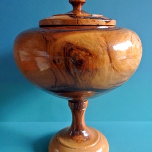 May include: A wooden bowl with a lid, on a pedestal base. The bowl is made of a dark wood with a natural grain pattern. The lid has a small knob on top.