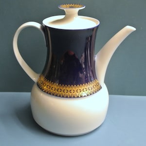 Puede incluir: Una tetera de porcelana blanca con un diseño azul y dorado. La tetera tiene un cuerpo redondo y un pico en el lateral. La tapa es blanca con un pomo dorado.