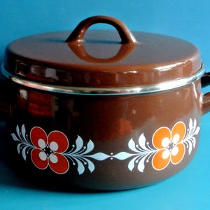 Puede incluir: Una olla de esmalte marrón con tapa y asas laterales. La olla presenta un diseño floral en rojo, naranja y blanco. La olla es probablemente para cocinar y tiene un acabado brillante.