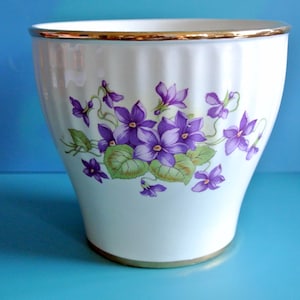 Könnte beinhalten: Weißer Keramiktopf mit goldfarbenem Rand und einem Blumendesign mit violetten und grünen Veilchen.