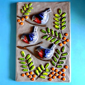 Puede incluir: Una placa de pared rectangular de cerámica con fondo marrón claro que presenta tres pájaros camachuelos en relieve posados sobre ramas marrones. El diseño incluye hojas verdes con reflejos amarillos y pequeñas bayas naranjas.