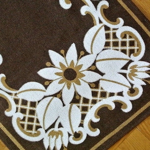 Könnte beinhalten: Ein braunes Stofftischset mit einem weißen und goldenen Blumendesign. Das Design zeigt stilisierte Blumen und Blätter mit einem Gittermuster im Hintergrund.