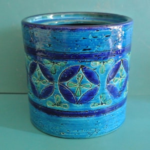 Puede incluir: Un jarrón cilíndrico de cerámica en tonos azules, con una superficie texturizada. El jarrón presenta un patrón repetitivo de círculos azul oscuro con diseños florales turquesa. El jarrón mide aproximadamente 18 cm de alto y 13 cm de diámetro.