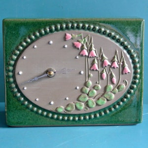 May include: A green ceramic clock with a white face and black hands. The clock is decorated with pink flowers and green leaves.